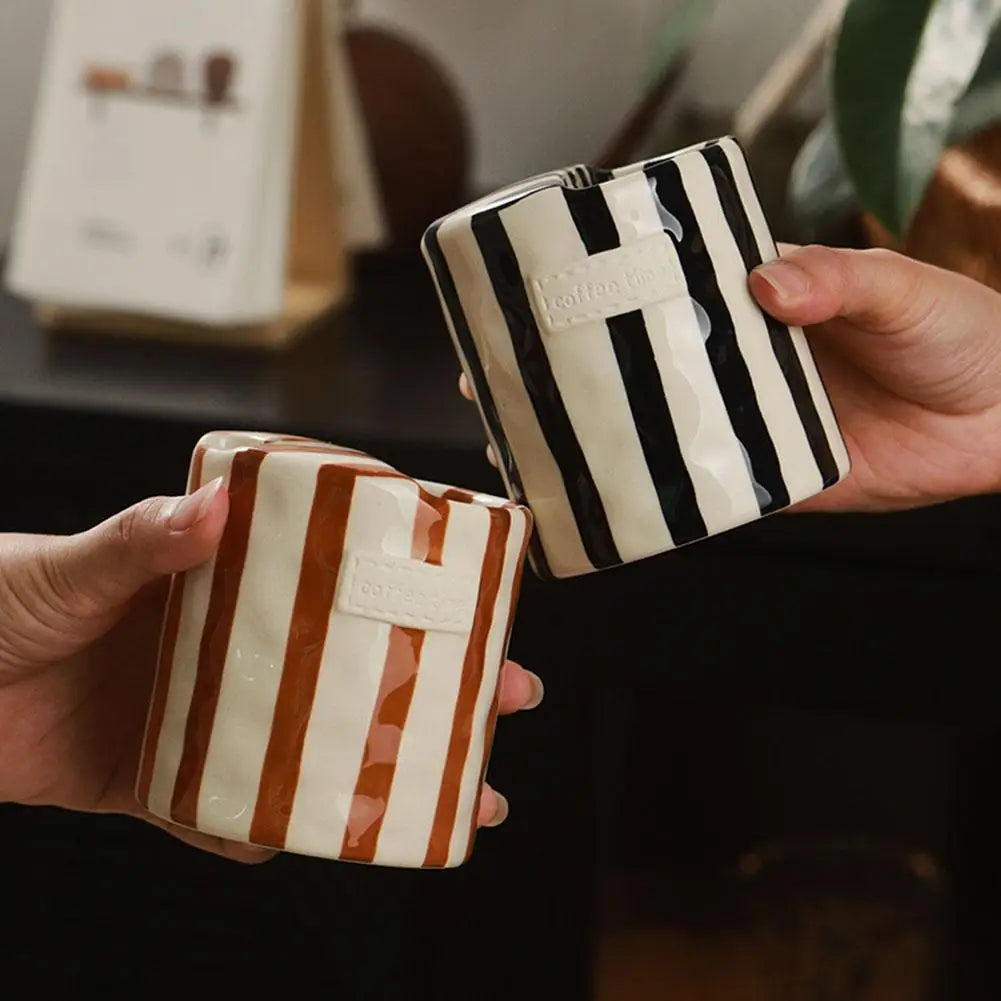 Two striped coffee mugs, one brown and white, the other black and white, held by hands against a blurred indoor background.