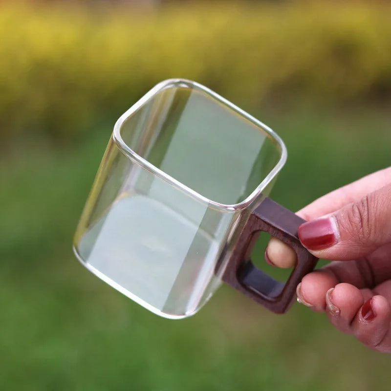 A person holding a minimalist square glass coffee cup by its ergonomic walnut wood handle against a soft green outdoor background.