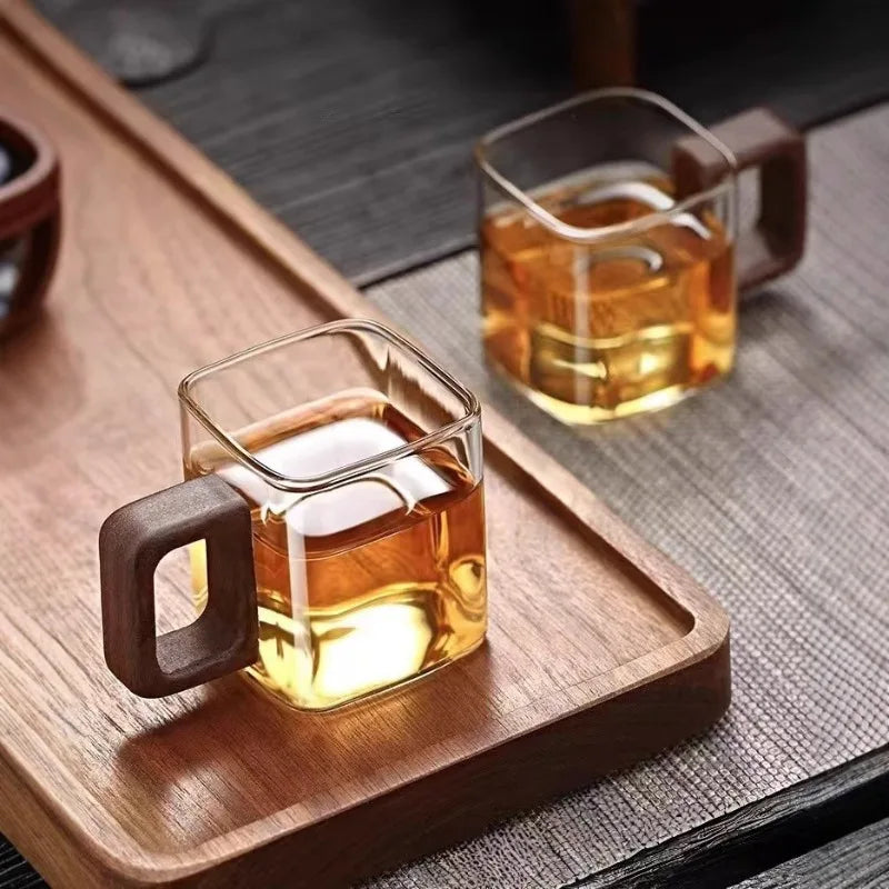 A pair of mid-century modern square glass mugs filled with warm tea on a sleek walnut wood tray.