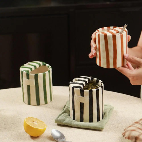 A lifestyle flat lay featuring two mid-century inspired striped ceramic mugs (one black/white, one green/brown) and a large black stripe mug with a hand holding a coffee bag on textured wooden tables.