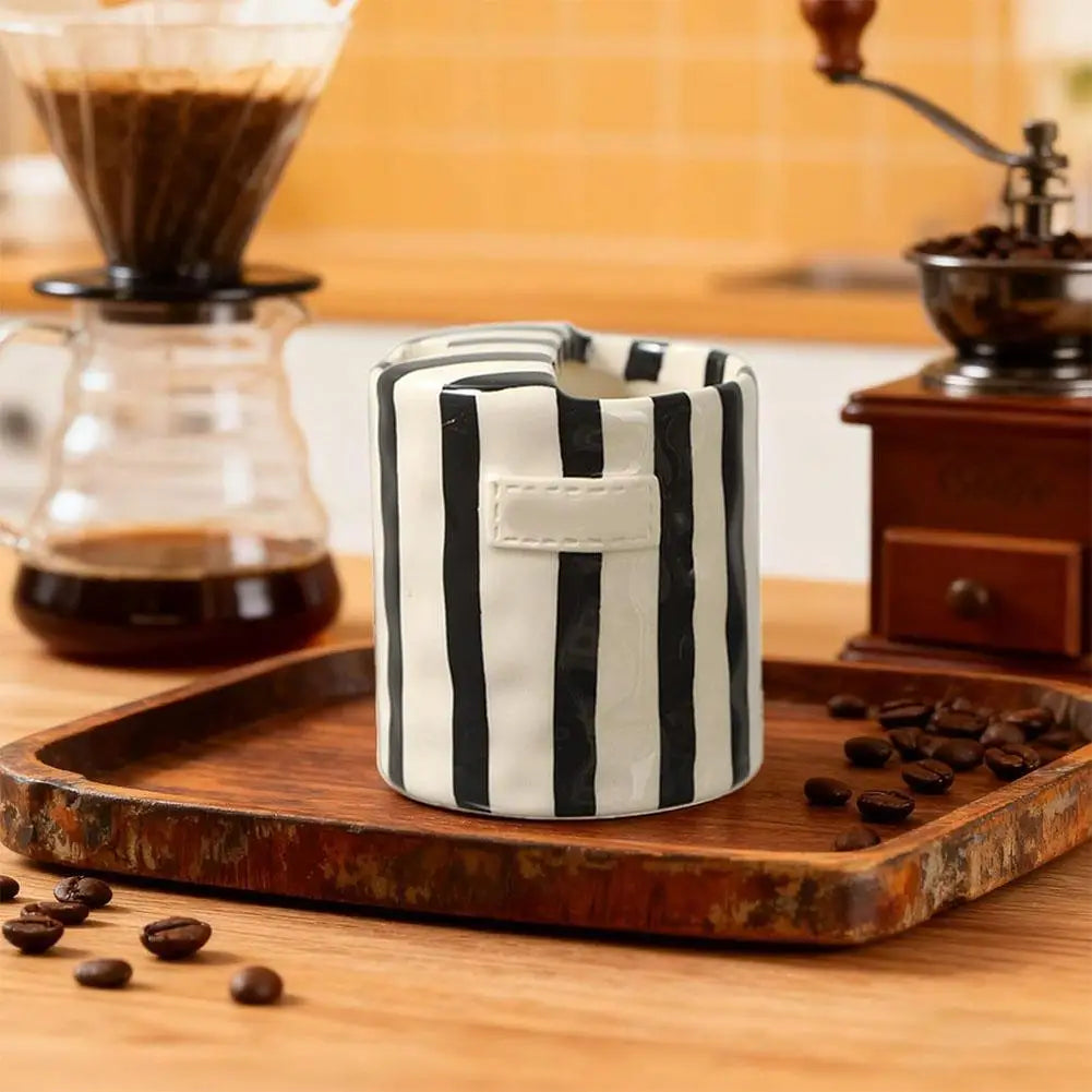 Black and white striped ceramic container on a wooden tray with coffee beans and a coffee grinder in the background.