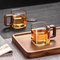 Two minimalist square glass coffee mugs with handcrafted walnut wood handles sitting on a modern wooden serving tray.